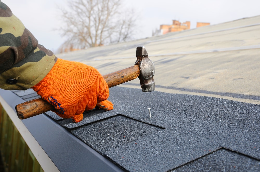 Worker Hands Installing Bitumen Roof Shingles Using Hammer In Nails.